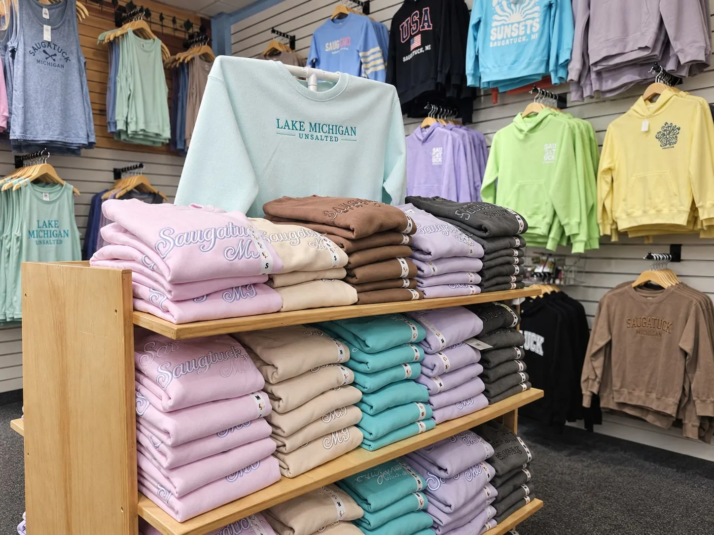 Lake Michigan Unsalted crewnecks and tanks beside folded Saugatuck script sweatshirts in pink, sand, teal and lavender