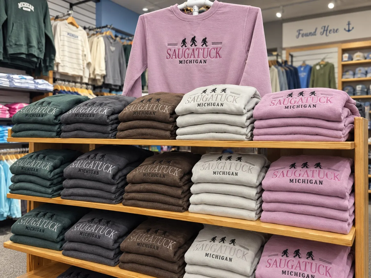 Folded Saugatuck Michigan crewnecks stacked on a wooden shelf in forest green, charcoal, brown, cream and pink
